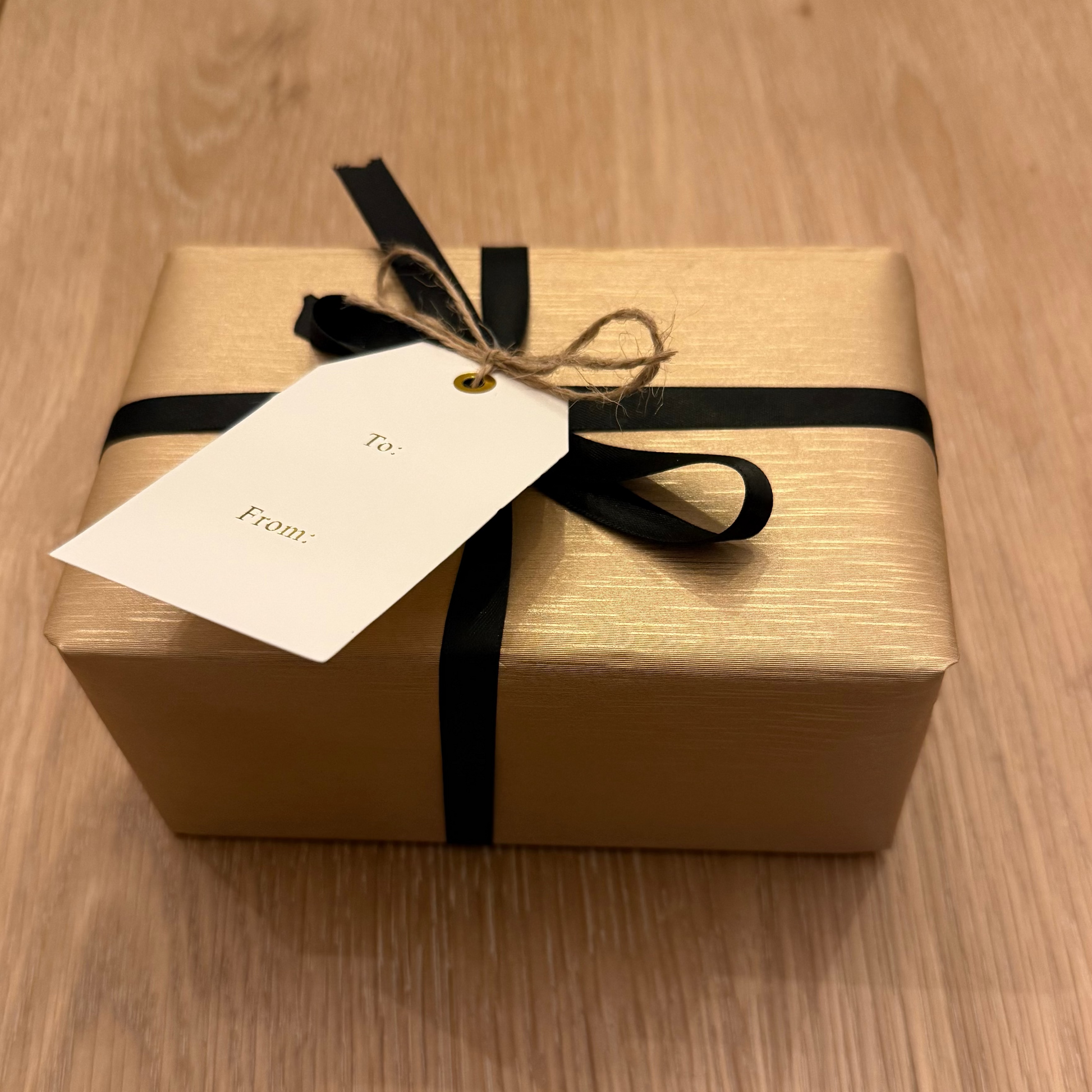 Gift box wrapped in textured, gold paper with a black ribbon and tag on a wooden surface