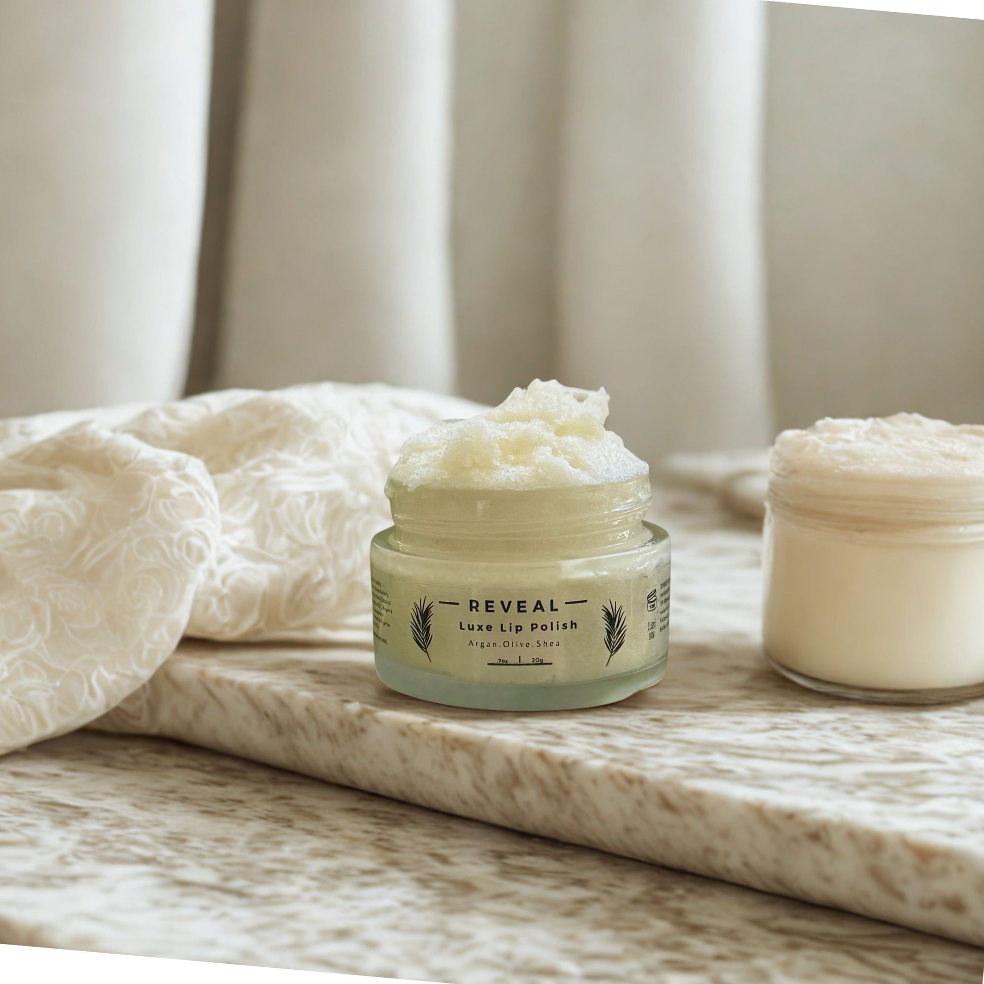 Jar of handmade creamy lip sugar scrub with hydrating oils and a natural color labeled 'REVEAL'  on a marble surface with a soft, neutral background. 